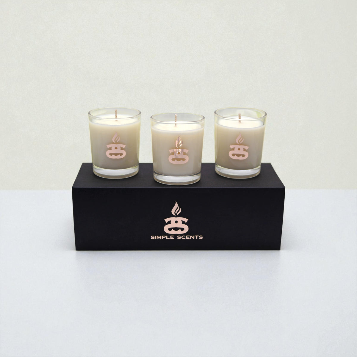 Simple Scents Mini Indulgence Trio Luxury Soy Candles 75g - three cream soy wax votive candles in glass jars with rose gold Simple Scents logo, displayed on top of matte black rectangular gift box on white background
