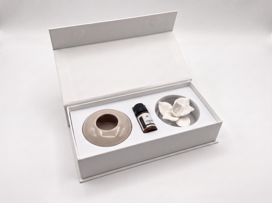 An open white gift box containing a beige ceramic diffuser base, a small dark bottle, and a white flower-shaped diffuser piece on a clean white background.