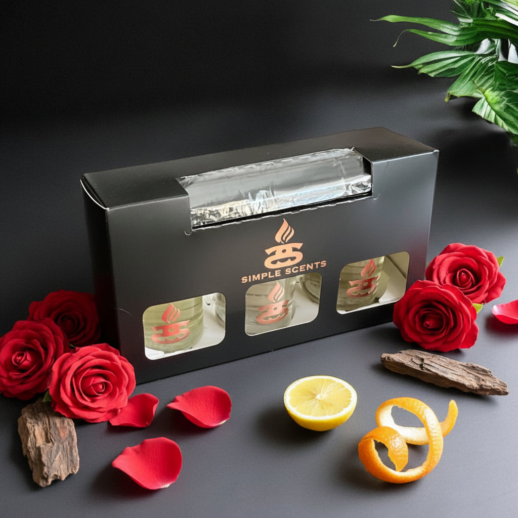 Simple Scents Excellence Reed Diffuser Trio Illicit Rose - black gift box with rose gold Simple Scents logo and three diffuser bottle windows, styled with deep red roses, rose petals, oud wood chunks, lemon half and orange peel curl on dark grey background