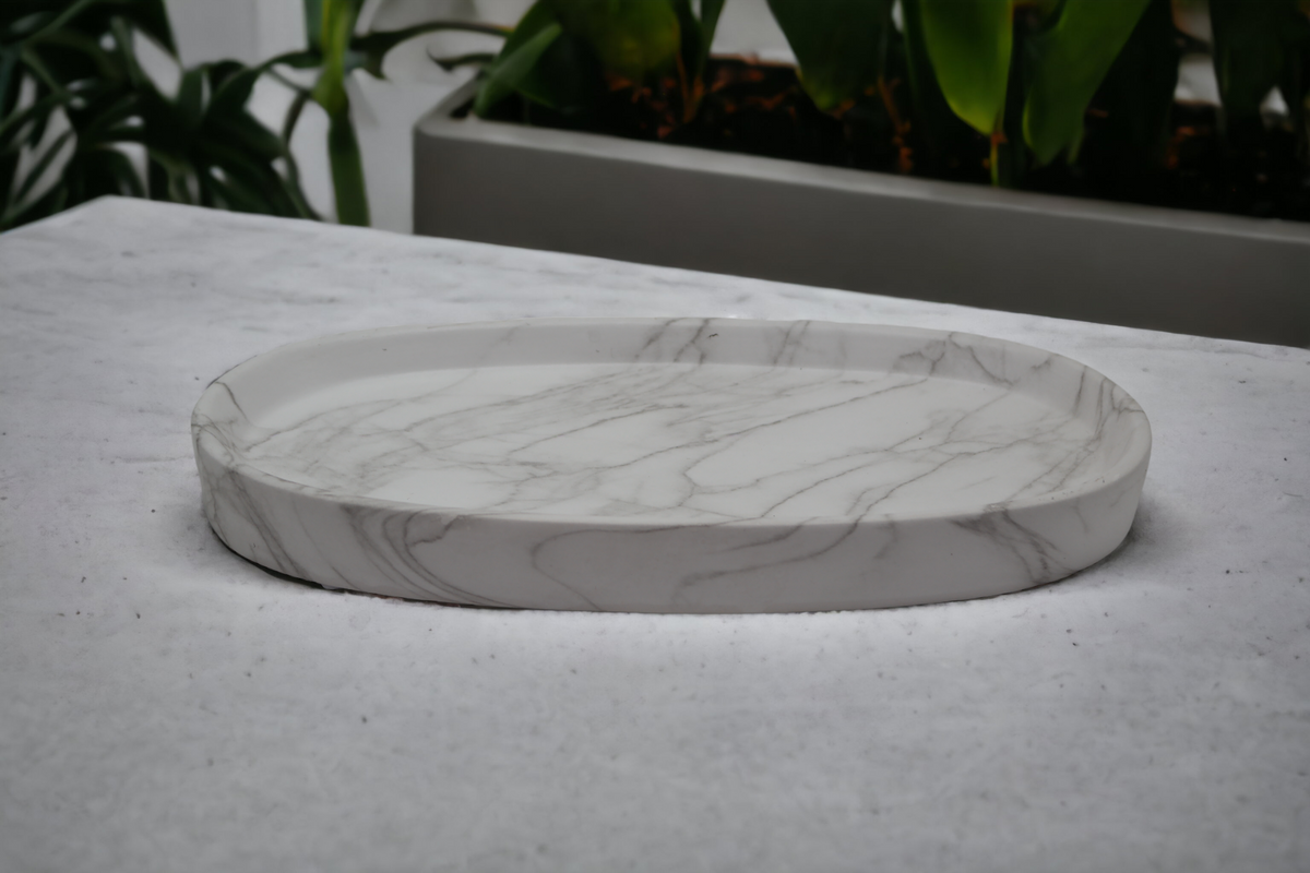 Oval Balmoral ceramic styling plate with grey marble-effect veining, displayed on a pale surface with green plants behind.