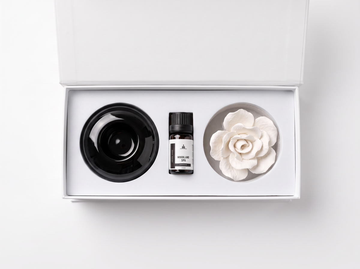 A white gift box containing a black diffuser bowl, a small bottle of essential oil, and a white flower-shaped diffuser on a clean white background.