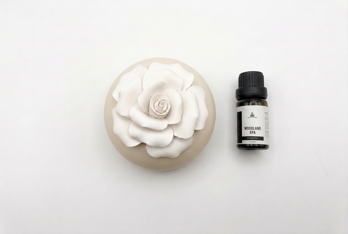 A round beige diffuser or decorative holder with a large white rose on top, shown next to a small black-and-white essential oil bottle labeled 'WOODLAND SPA' on a plain white background.