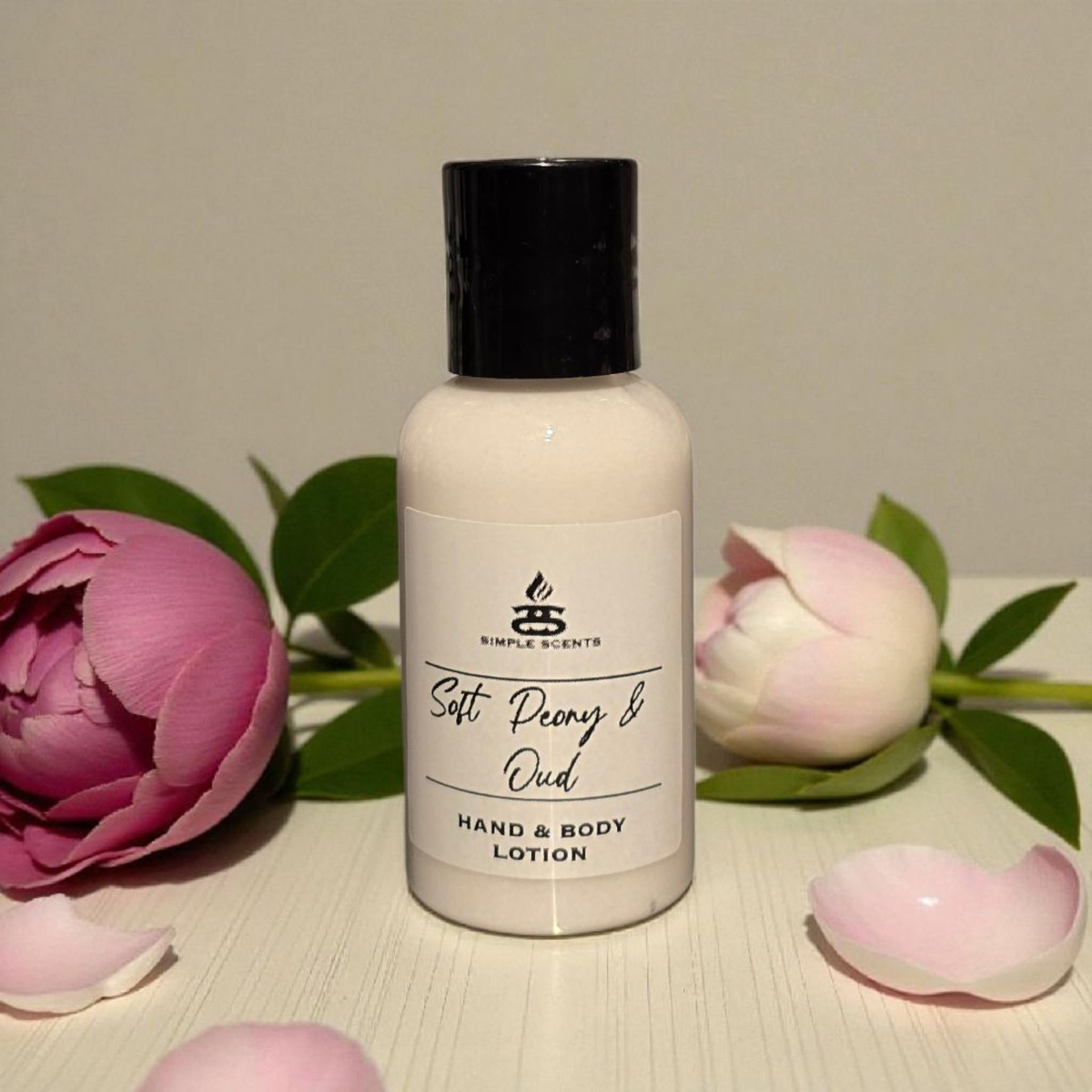Simple Scents Minis Soft Peony & Oud Hand & Body Lotion 50ml - small white bottle with black cap and Simple Scents label, styled on white wooden surface flanked by a deep pink peony bloom, a pale pink peony bud and scattered pink petals
