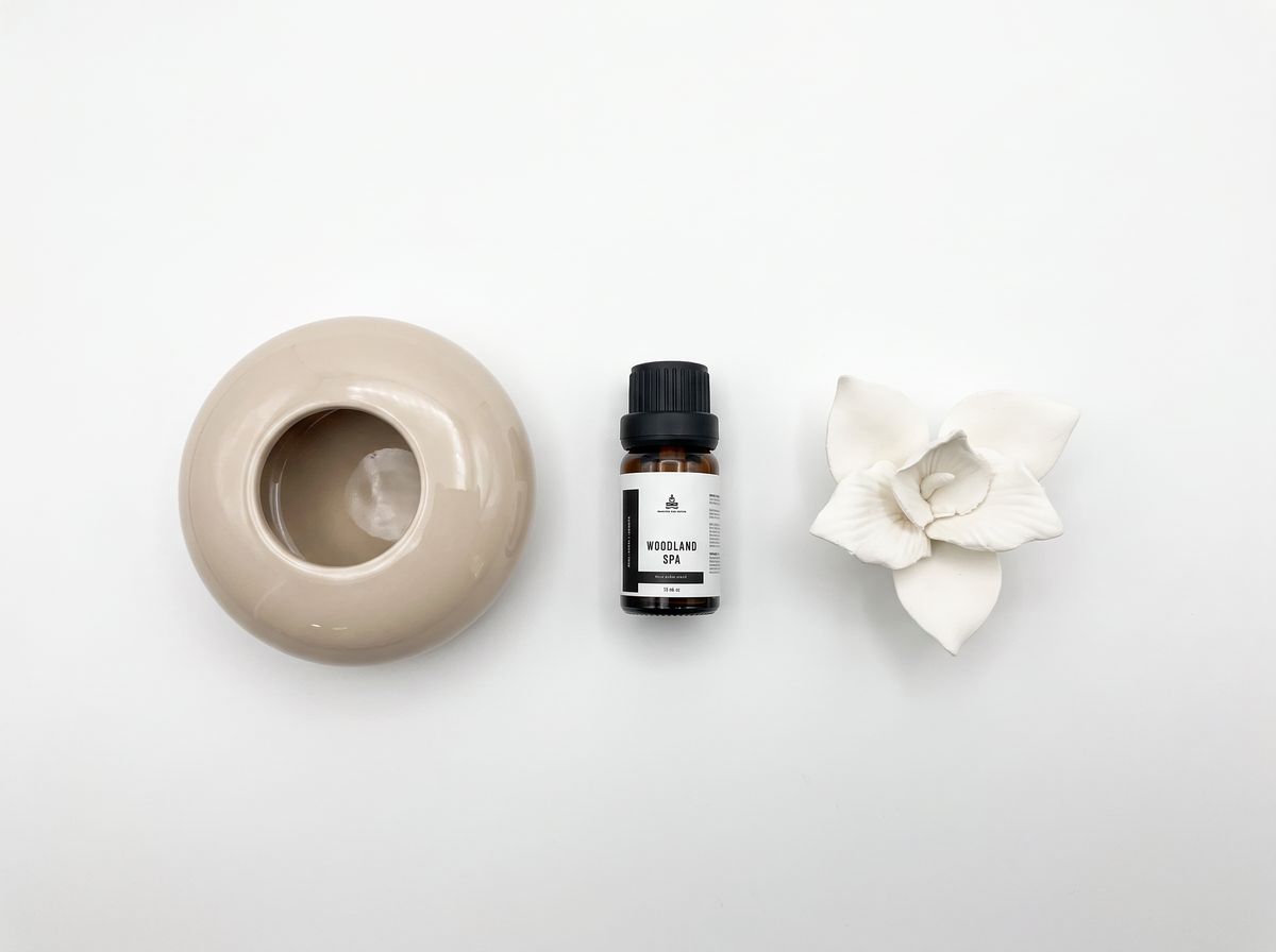 A small amber glass bottle with a black cap and white label sits centered between a beige ceramic diffuser bowl on the left and a white flower-shaped diffuser on the right, all arranged on a clean white background.