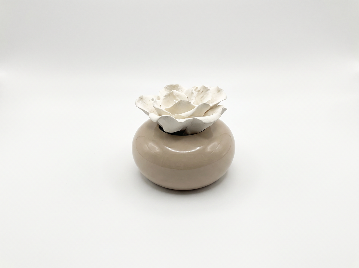 A small ceramic container with a smooth, rounded beige base and a white sculpted flower-shaped diffuser stone, set against a seamless light grey studio background.