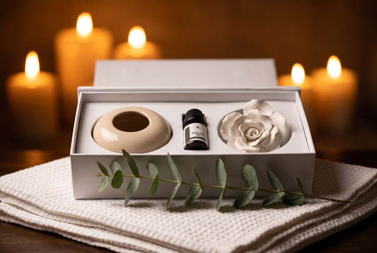 A gift box containing a beige ceramic bowl, a small black bottle, and a white ceramic rose, styled on a textured cream towel with eucalyptus leaves and glowing candles in the background.