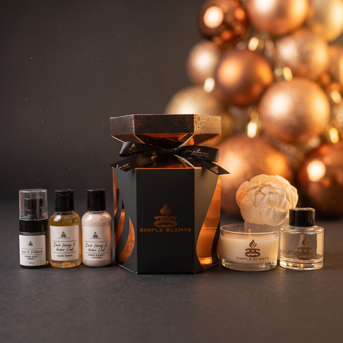 A Simple Scents gift set featuring a black and copper gift box with a black ribbon, three small travel-sized bottles, a cream peony flower-shaped sola reed, and a clear glass diffuser, arranged on a dark surface with a blurred background of metallic copper ornaments.