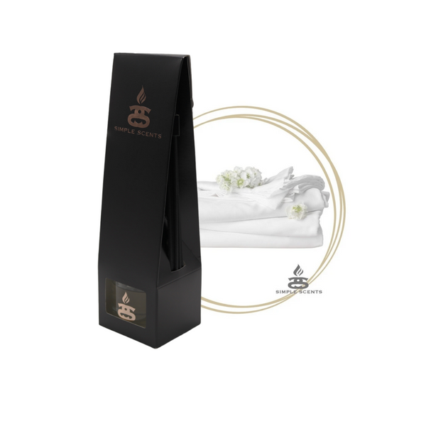 Fresh Linen Simple Scents Excellence Reed Diffuser - matte black tall tapered packaging box with rose gold Simple Scents logo, displayed in front of neatly folded white linen and small white flowers inside a gold double-ring circle frame on white background