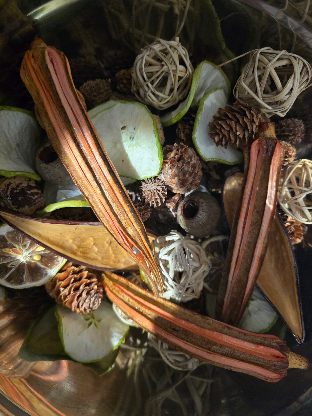 A close-up of a decorative dried botanical arrangement in a glass container, featuring reddish-brown seed pods, pinecones, light green apple slices, and white woven ball ornaments.