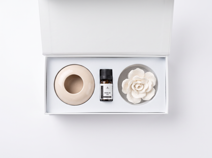 A diffuser gift set in a white box, featuring a beige ceramic diffuser base, a small amber bottle with a black cap, and a white ceramic flower diffuser.