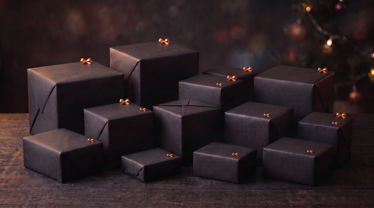 Gifts in assorted sizes wrapped in black paper with small copper bows, arranged on a wooden table beside softly glowing. lights.