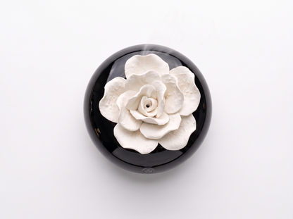 A round black object with a raised white flower design on top, photographed from above on a plain white background.