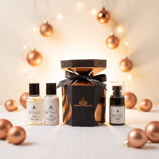 A gift set of Simple Scents hand and body products arranged on a light surface with decorative rose-gold ornaments and warm fairy lights in the background. The set includes a black and gold gift box tied with a black ribbon, two small clear bottles with black caps, and one larger clear bottle with a black pump.