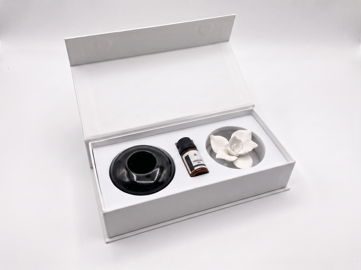A white gift box with its lid open, containing a black circular diffuser base, a small amber bottle with a white label, and a white flower-shaped diffuser piece on a white insert.