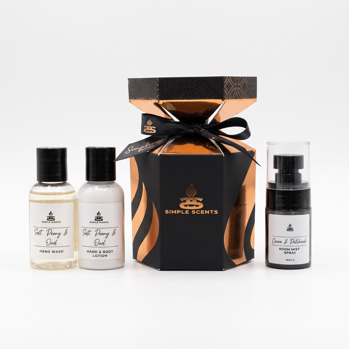 A Simple Scents gift set containing a central black and gold hexagonal box tied with a black ribbon, flanked by two small bottles of hand wash and body lotion on the left, and a pump bottle of room body spray on the right, all on a white background.