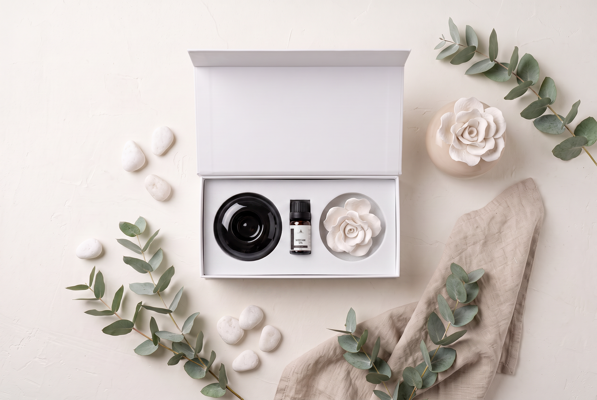An open white gift box on a light beige surface, containing a black round container and a small black bottle with a white label, alongside a white flower-shaped item. The scene is styled with eucalyptus branches, white stones, a beige cloth, and a ceramic flower vase.