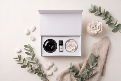 An open white gift box on a light beige surface, containing a black round container and a small black bottle with a white label, alongside a white flower-shaped item. The scene is styled with eucalyptus branches, white stones, a beige cloth, and a ceramic flower vase.