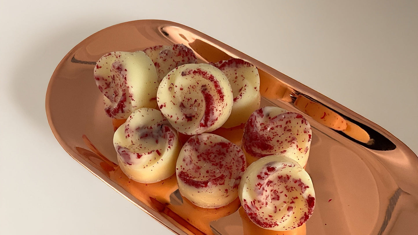 A set of white wax melt swirls with red pomegranate flower sprinkles, arranged on a reflective copper tray against a neutral light gray background.