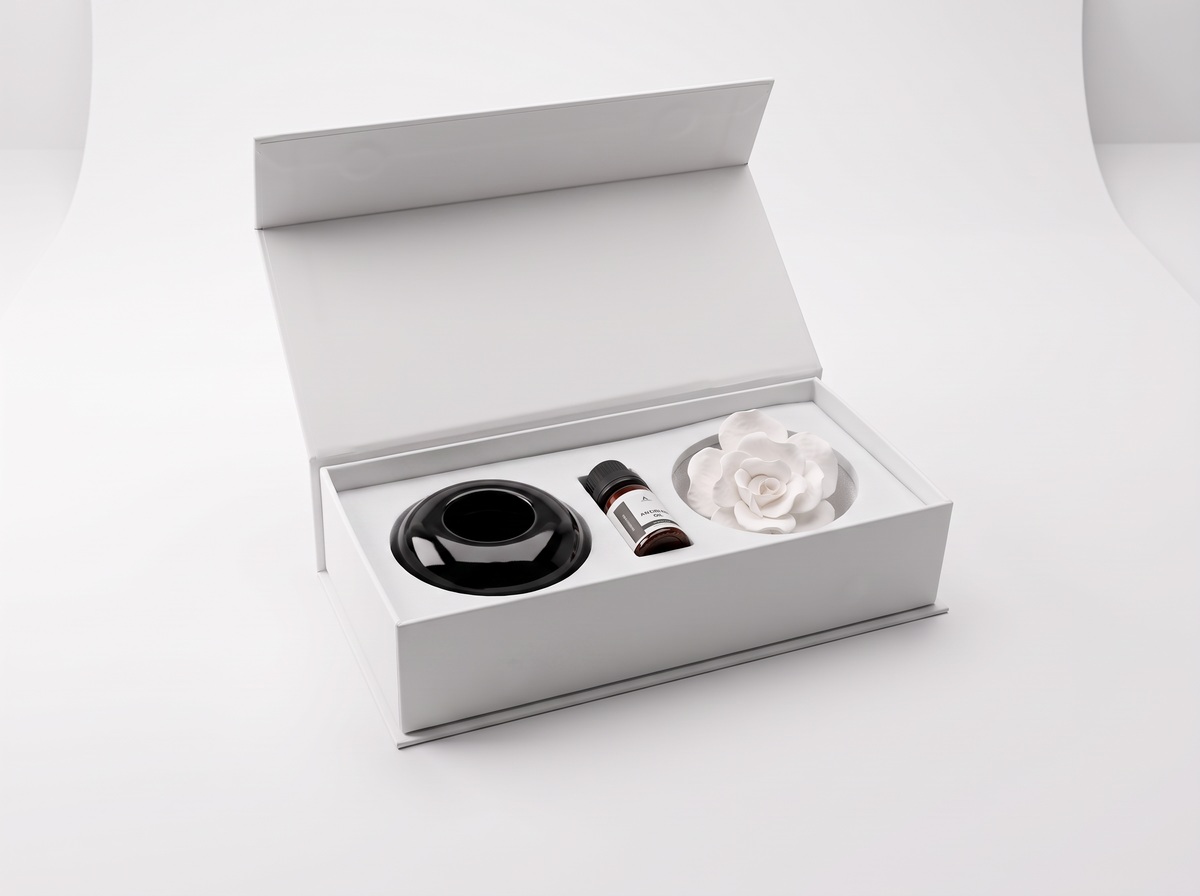 An open white gift box on a clean white background, containing a black circular diffuser base, a small amber bottle, and a white rose-shaped diffuser.