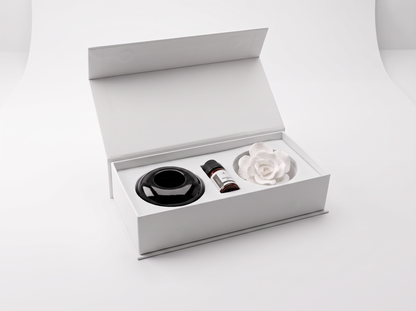 An open white gift box on a clean white background, containing a black circular diffuser base, a small amber bottle, and a white rose-shaped diffuser.