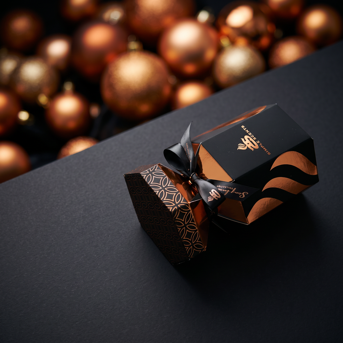 A luxury gift box in matte black with metallic copper-gold geometric patterns and a black ribbon bow, set on a dark surface with a background of blurred copper Christmas ornaments.