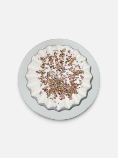 Simple Scents Tea Tree and Rosemary Bath Bomb with scattered dried floral botanicals, viewed from above on a light grey background