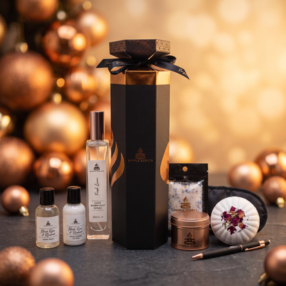 A luxury gift set from Simple Scents arranged on a dark surface with a festive blurred background of gold Christmas ornaments. The set includes a tall black and copper gift box with a black ribbon, a clear bottle of room spray, two small bottles of hand wash and lotion, a tin candle, a sachet of bath salts, a black eye mask, and a decorative pen.