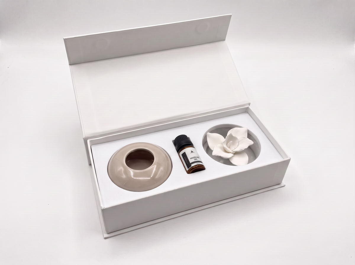 An open white gift box containing a beige ceramic diffuser base, a small dark bottle, and a white flower-shaped diffuser piece on a clean white background.