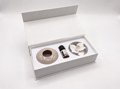 An open white gift box containing a beige ceramic diffuser base, a small dark bottle, and a white flower-shaped diffuser piece on a clean white background.