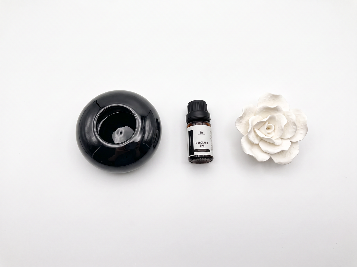 A diffuser set consisting of a black round ceramic diffuser bowl, a small amber bottle of essential oil, and a white ceramic flower, arranged on a clean white background.