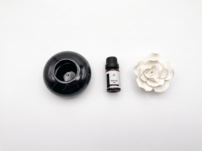 A diffuser set consisting of a black round ceramic diffuser bowl, a small amber bottle of essential oil, and a white ceramic flower, arranged on a clean white background.