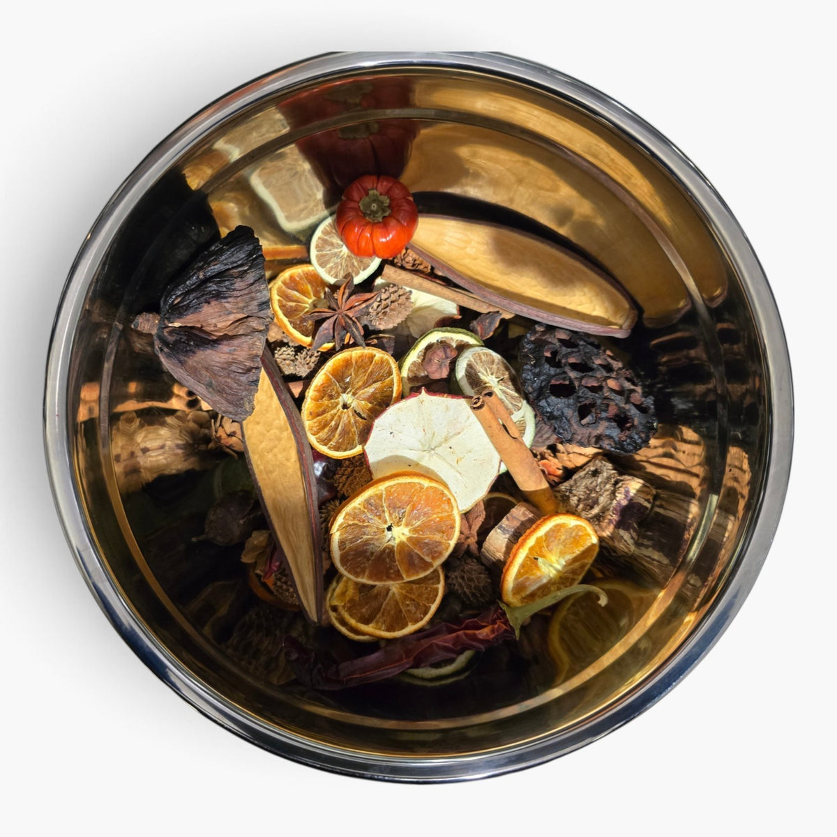 A top-down view of a round stainless steel pot filled with dried botanicals and citrus slices, including orange slices, cinnamon sticks, star anise, a small dried tomato, and a dark brown dried flower, set against a plain white background.