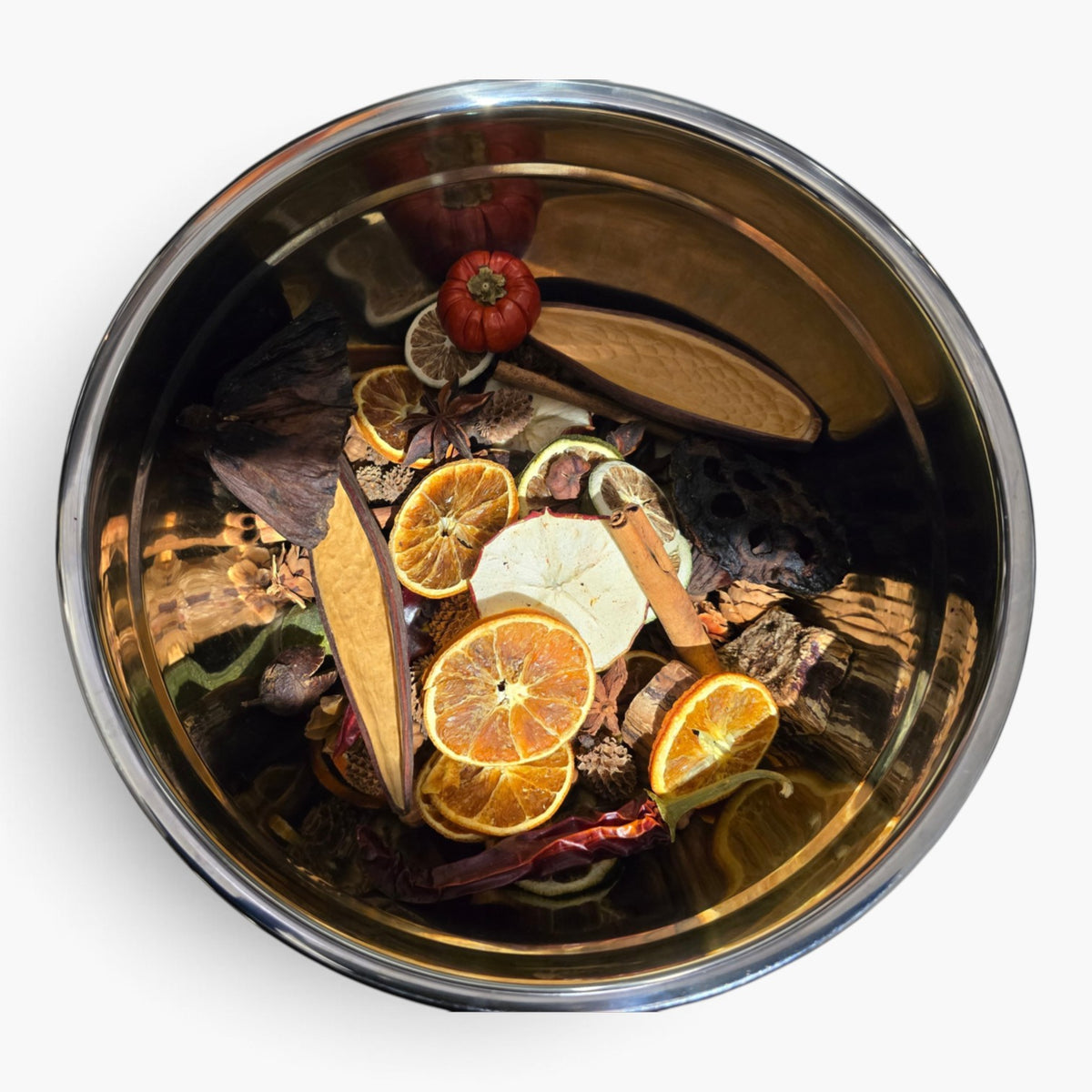 A top-down view of a stainless steel bowl filled with dried botanicals and citrus slices, including orange, cinnamon, and a small red dried fruit, on a plain white background.