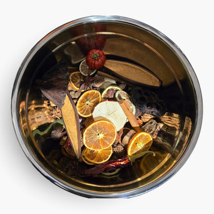 A top-down view of a stainless steel bowl filled with dried botanicals and citrus slices, including orange, cinnamon, and a small red dried fruit, on a plain white background.