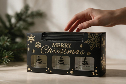 Hand reaching towards a Simple Scents Christmas reed diffuser trio gift box on a wooden table beside evergreen branches.