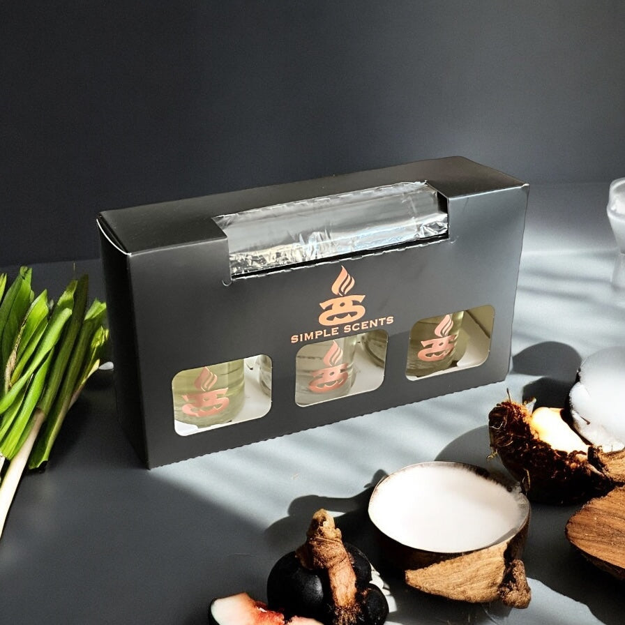 Simple Scents Excellence Reed Diffuser Trio Essence - black gift box with rose gold Simple Scents logo and three diffuser bottle windows, styled with lemongrass stalks, fresh ginger root, coconut halves and galangal on dark grey background