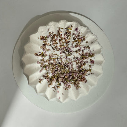 Overhead close-up of Simple Scents Tea Tree and Rosemary Bath Bomb with dried purple heather flower botanicals on a grey background