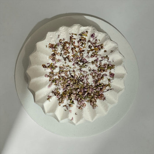 Overhead close-up of Simple Scents Tea Tree and Rosemary Bath Bomb with dried purple heather flower botanicals on a grey background