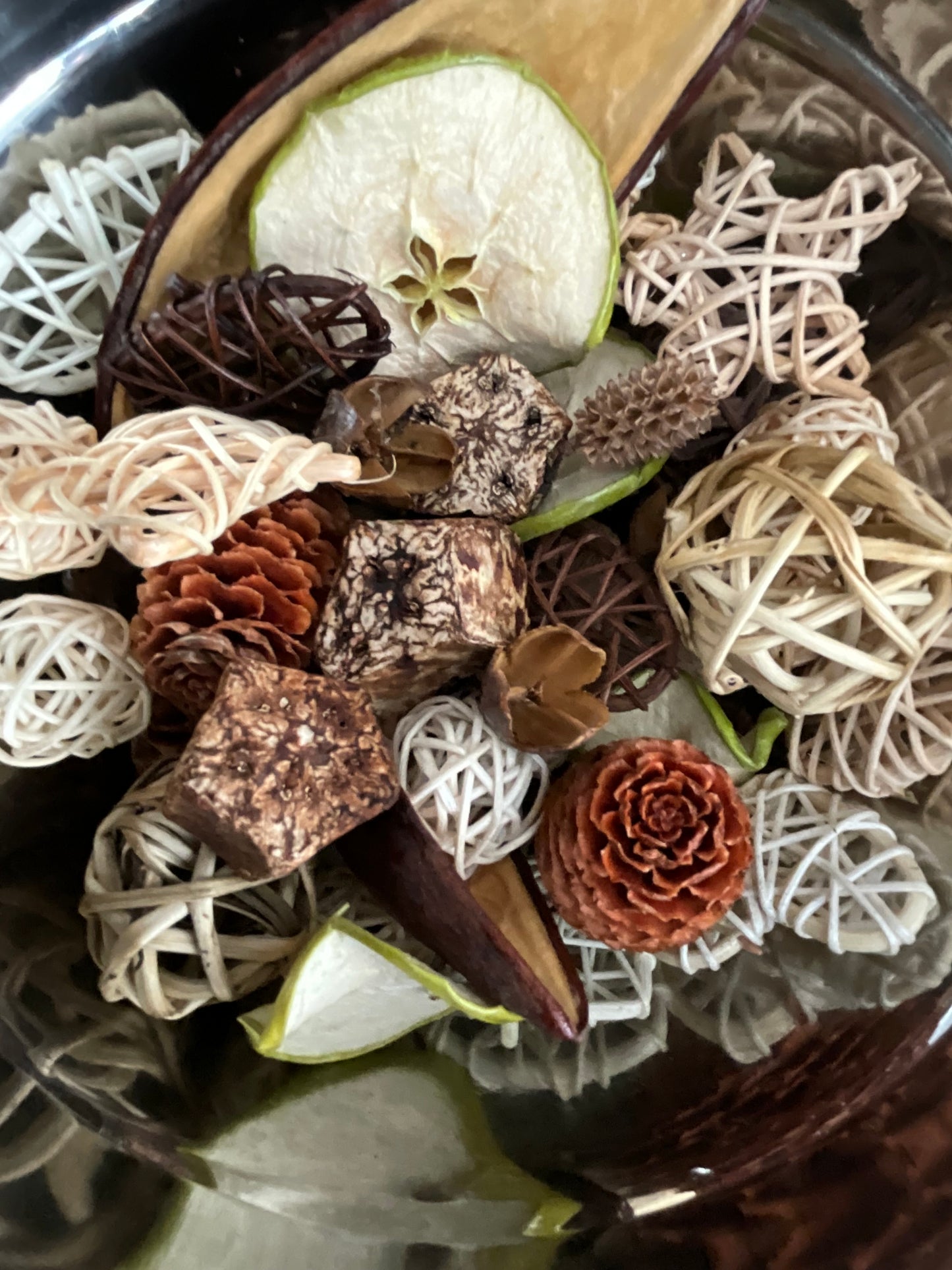 A close-up of a decorative dried botanical arrangement in a dark bowl, featuring woven rattan balls, pinecones, bark pieces, and sliced green apple.