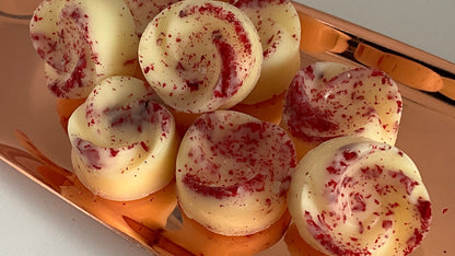 Several round wax melts with a white base and red swirl pattern, arranged on a metallic copper tray against a plain white background.
