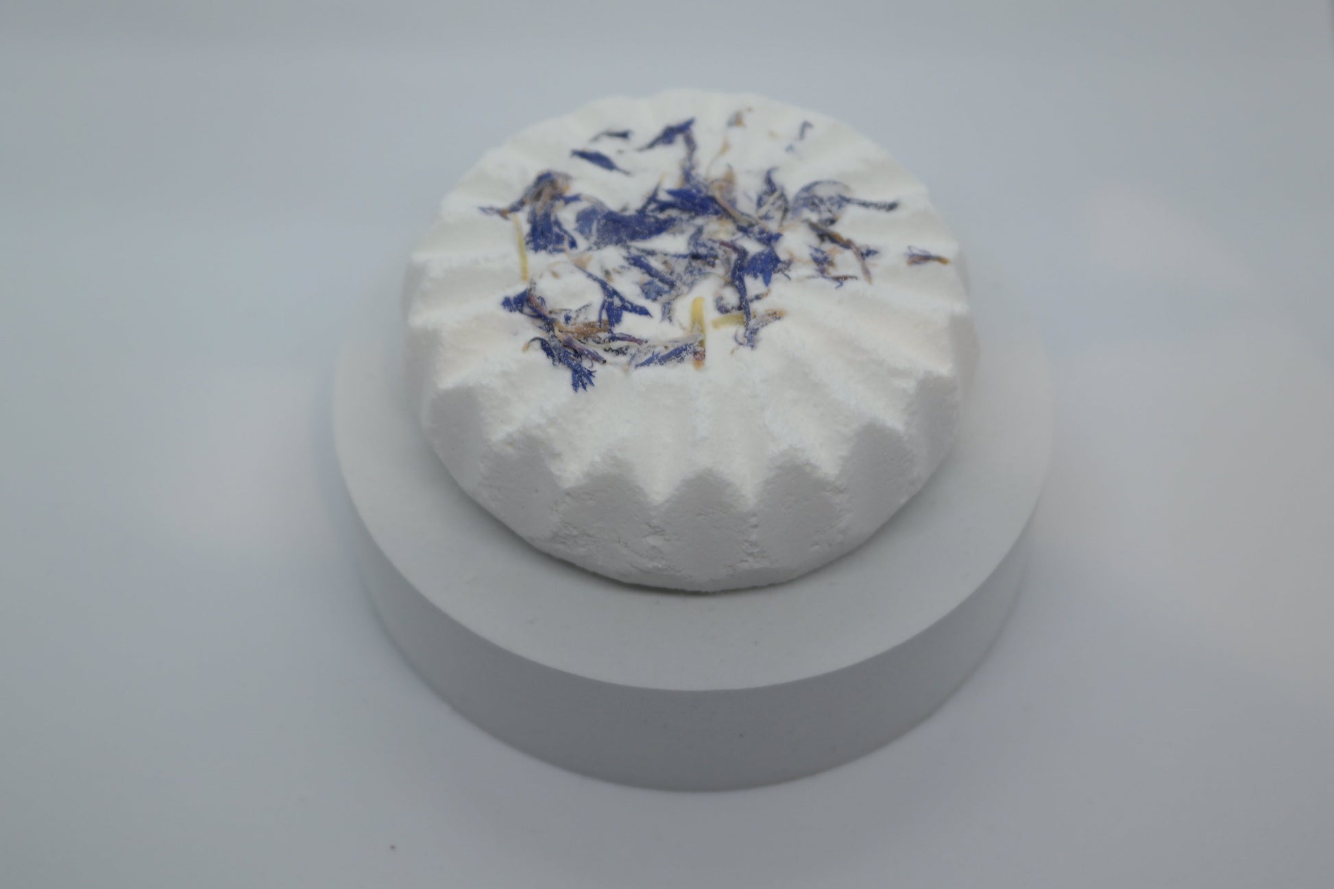 Simple Scents Aromatherapy Lavender & Frankincense Bath Bomb with dried blue cornflower petals, displayed on a white round plinth against a light grey background