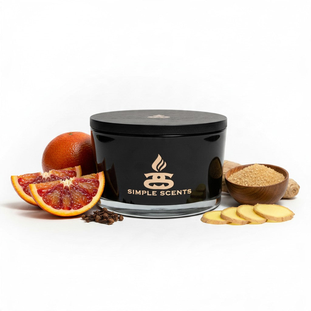 A black glass candle jar with a black lid and gold 'SIMPLE SCENTS' logo, styled with citrus slices, ginger slices, cinnamon sticks, and a small bowl of wax on a white background.