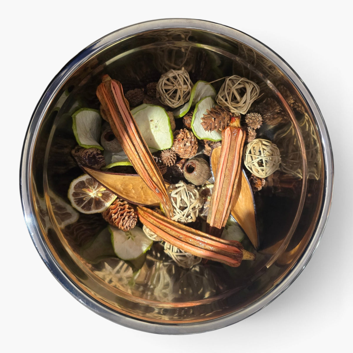 A top-down view of a clear glass bowl filled with dried botanicals, including cinnamon sticks, pinecones, small woven balls, and dried citrus slices, on a white background.