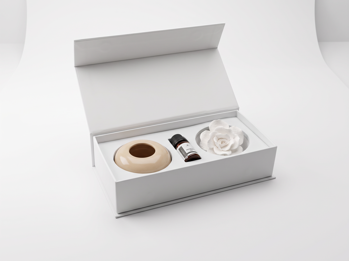 An open white gift box containing a beige ceramic diffuser, a small dark bottle, and a white flower-shaped diffuser insert, photographed on a clean white background.