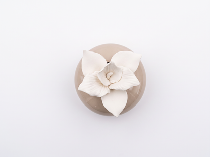 A small round beige ceramic dish with a glossy finish, topped with a sculpted white flower with layered petals, photographed from directly above on a seamless white background.