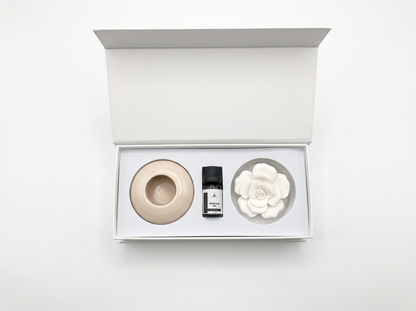 An open white gift box containing a minimalist three-piece set: a beige ceramic diffuser bowl, a small black-and-white labelled bottle, and a white ceramic flower-shaped diffuser, set against a clean white background.