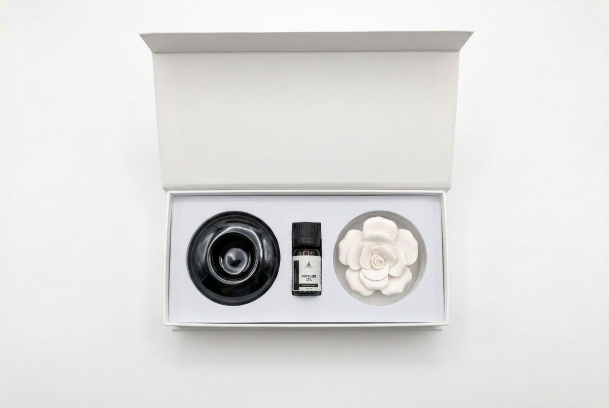An open white gift box containing a black circular diffuser, a small amber bottle, and a white flower-shaped diffuser on a clean white background.