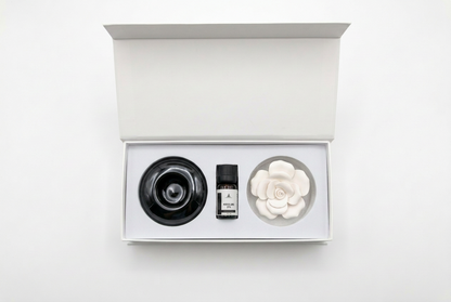 An open white gift box containing a black circular diffuser, a small amber bottle, and a white flower-shaped diffuser on a clean white background.