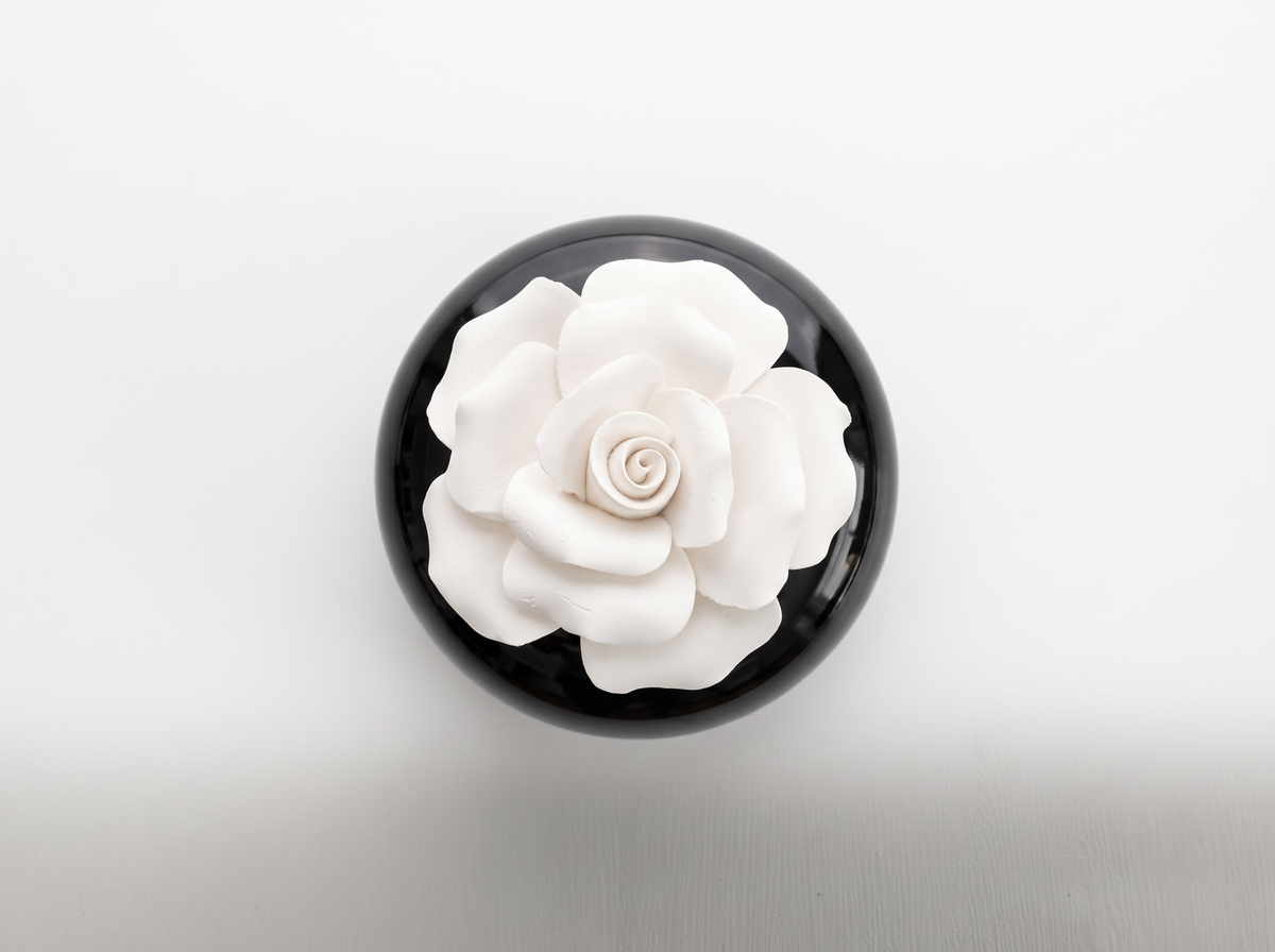 A round black container or box viewed from above, featuring a large white sculpted flower on the lid, set against a clean white background.