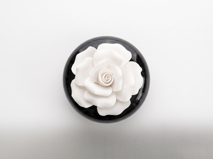 A round black container or box viewed from above, featuring a large white sculpted flower on the lid, set against a clean white background.